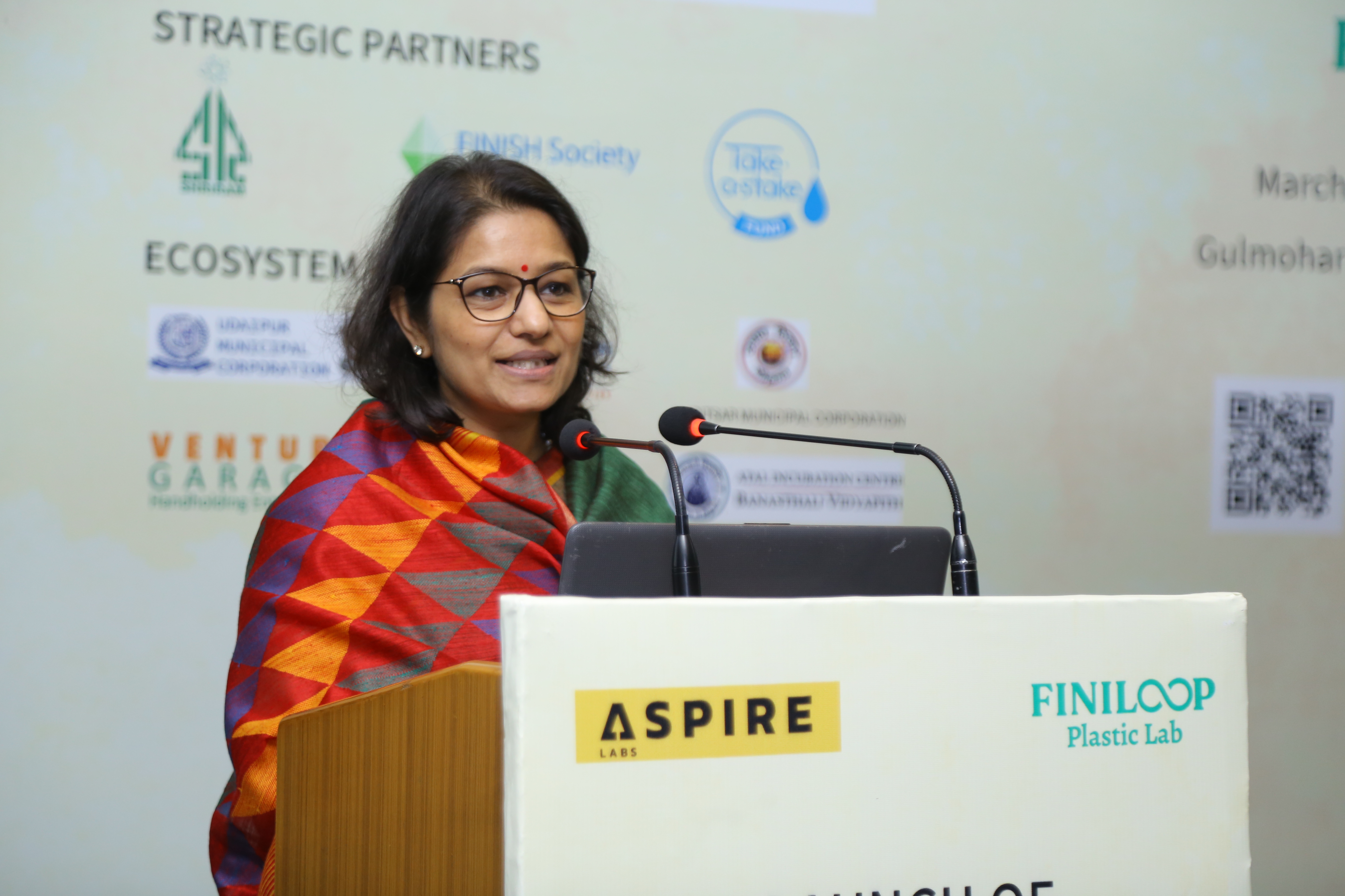 Ms. Saloni Goel in a red scarf presenting from a podium at the Aspire Labs event 