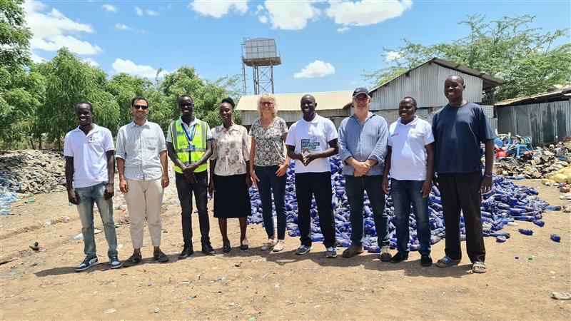 9 members of the SWEEP Partnership stood smiling in a row in front of a pile of blue plastic bottles at Kakuma Refugee Camp