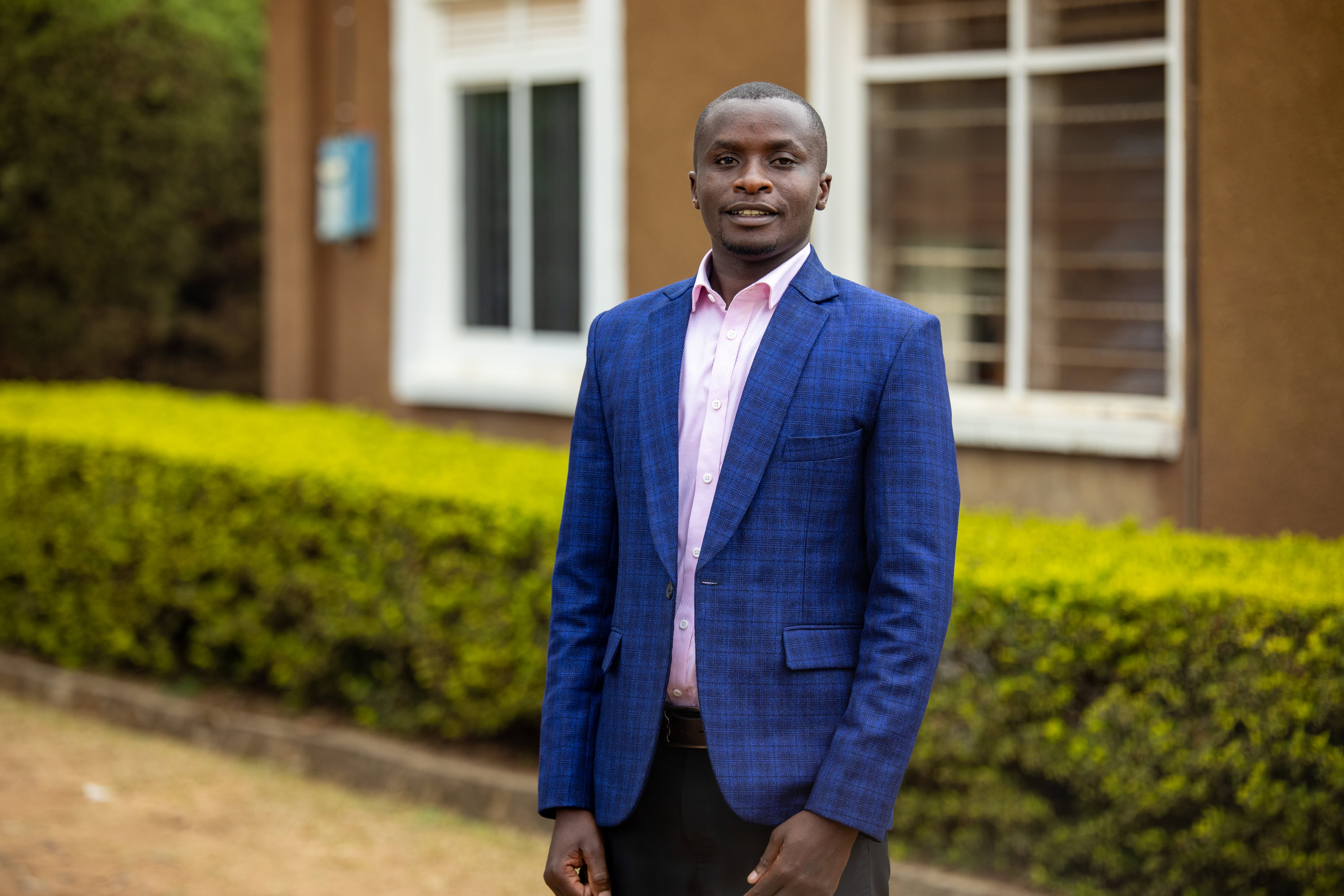 Denison in a blue suit standing up in front of a green hedge and brown building 